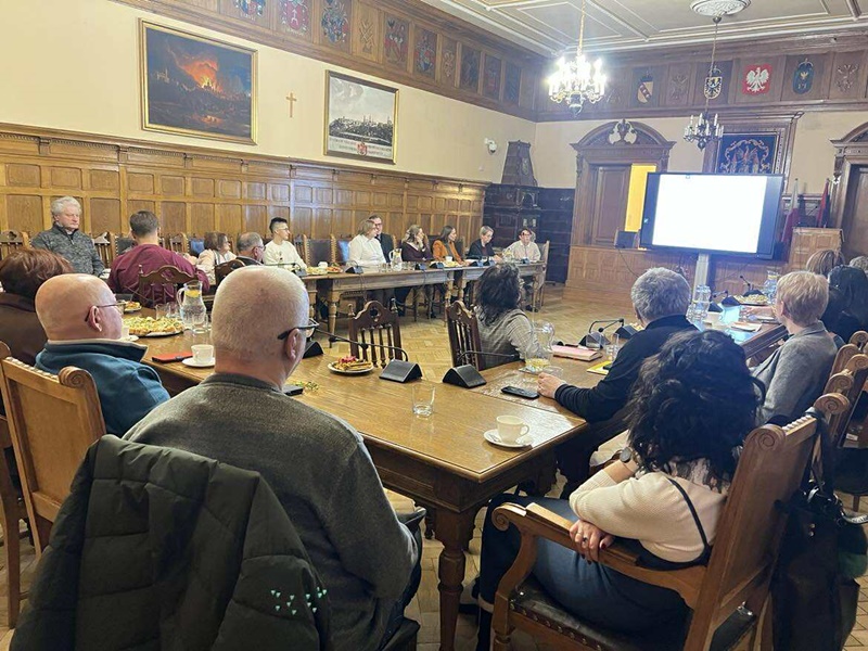 Spotkanie Noworoczne z udziałem przedstawicieli organizacji pozarządowych w Sali Sesyjnej cieszyńskiego Ratusza. Duża, zabytkowa sala. Przy długim stole siedzi kilkanaście osób i patrzy na ekran z prezentacją. Na stole są mikrofony, woda i filiżanki. 